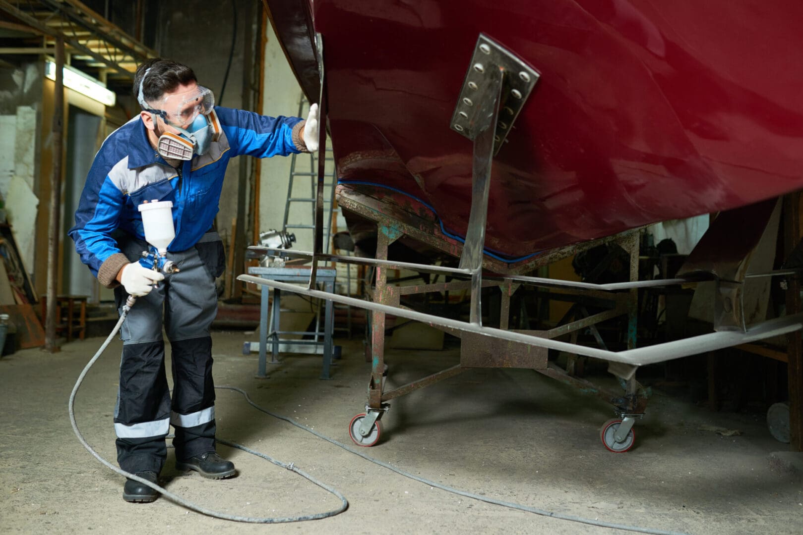 Full length portrait of  worker wearing protective mask checking coating while painting boat in yacht workshop using paint sprayer, copy space