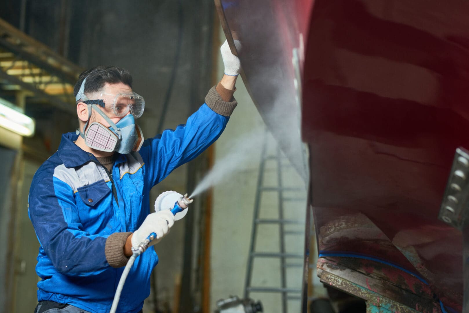 Side view portrait of  worker wearing protective mask spray painting boat in yacht workshop  copy space